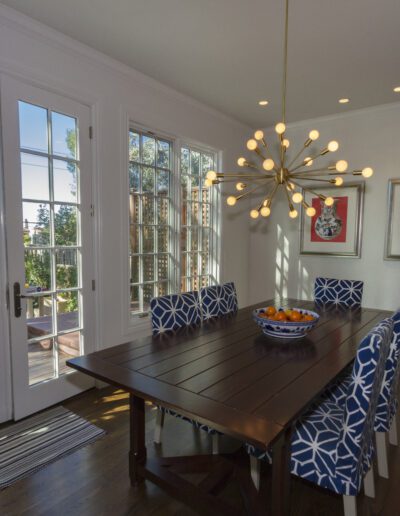 A bright dining room with a wooden table, blue patterned chairs, starburst chandelier, and artwork on the walls, opening onto a balcony through french doors.