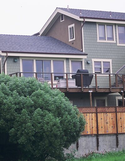 A two-story green house with an exterior staircase and a wooden fence surrounded by greenery.