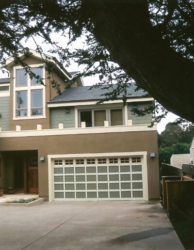 A two-story residential home with a beige facade, a large garage door, and several windows, surrounded by trees and a wooden fence.