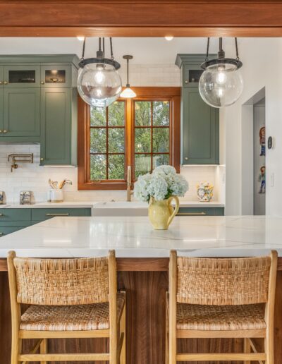 Modern kitchen with teal cabinetry, white countertops, rattan bar stools, and pendant lights, viewed from an adjacent room with wooden trim.