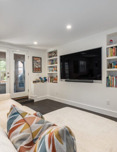 A modern living room featuring a large couch with colorful pillows, mounted tv, bookshelves, artworks, and glass doors leading outside.