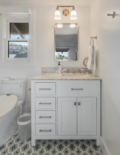 A clean, modern bathroom featuring a white vanity with drawers and cabinets, a wall-mounted mirror, overhead lighting, and a patterned tile floor.