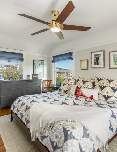 Bright, airy bedroom with a floral bedspread, wooden floor, ceiling fan, and various artworks on walls, with a view of a cityscape through multiple windows.