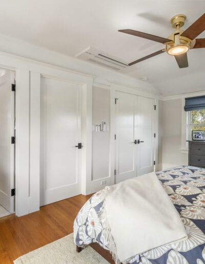 A bright, modern bedroom featuring a ceiling fan, large bed with patterned cover, built-in white closets, and an adjoining bathroom with marble walls.